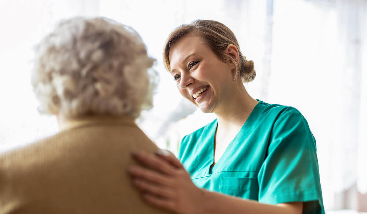 Carer in green scrubs assisting elderly woman with grey hair